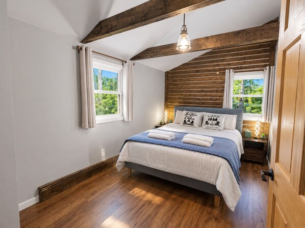 Une chambre avec un plafond en bois, un lit recouvert d’une courtepointe bleue et blanche, deux lampes de chevet et une fenêtre donnant sur un paysage verdoyant.