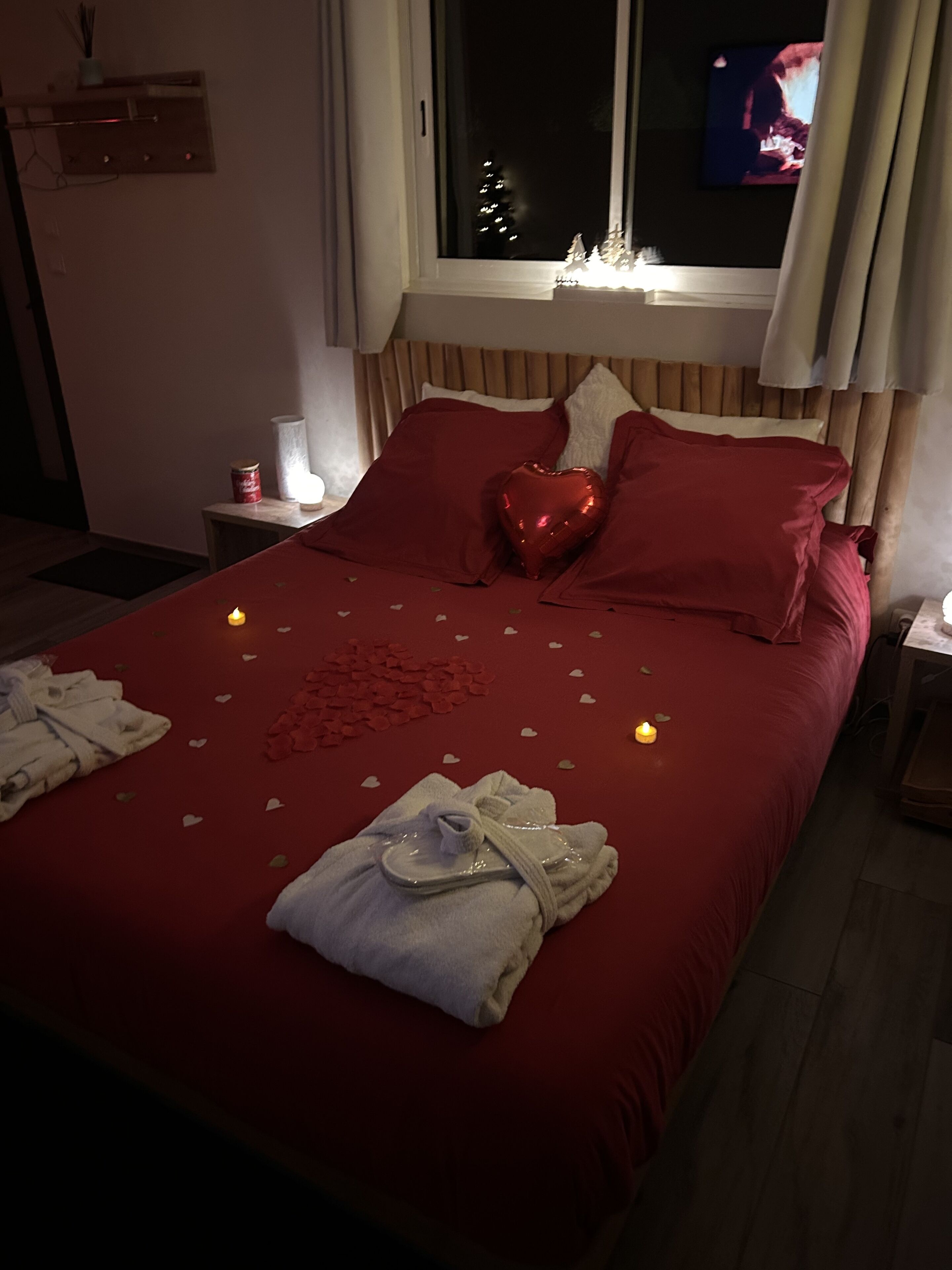 Un lit bien fait, avec une literie rouge, des oreillers en forme de cœur et une décoration en forme de cœur. On y voit des bougies et une petite table placée à côté.