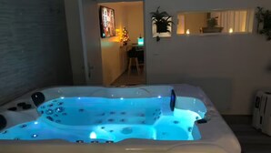 Un jacuzzi con iluminación azul, un acceso a una habitación con escritorio y silla, y un ventanal con plantas.