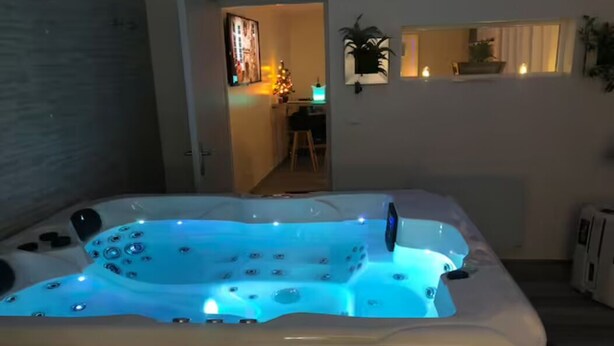 Un jacuzzi con iluminación azul, un acceso a una habitación con escritorio y silla, y un ventanal con plantas.