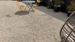 Un patio con una mesa redonda y dos sillas amarillas, rodeado de plantas en macetas y una cerca negra.