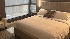 Quarto moderno com uma cama grande, uma mesa de cabeceira, uma cadeira e uma janela com cortinas leves.