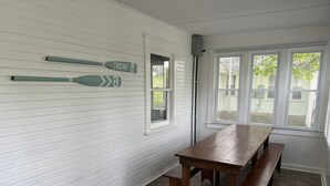 Une pièce avec une table et des bancs en bois, des murs recouverts de panneaux blancs et deux fenêtres équipées de stores.