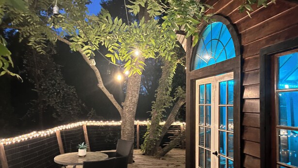A wooden deck with a table and chairs, surrounded by string lights and a tree. - Cozy Treehouse on Lake Conroe w/ large nest deck with a creek view (Willis)