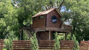 A wooden treehouse with a circular window, built on stilts, surrounded by benches and trees. - Cozy Treehouse on Lake Conroe w/ large nest deck with a creek view (Willis)