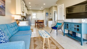 A spacious living area with a blue sofa, a coffee table, and a television. There is a kitchen in the background with white cabinets and a dining area with a table and chairs. - Bayfront Pensacola Beach Condo w/ Pool & Elevator (Pensacola Beach)