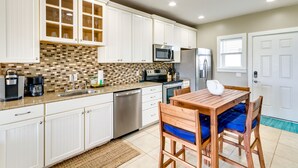 A kitchen with white cabinets, a tiled backsplash, stainless steel appliances, and a wooden dining table with blue cushions. - Bayfront Pensacola Beach Condo w/ Pool & Elevator (Pensacola Beach)