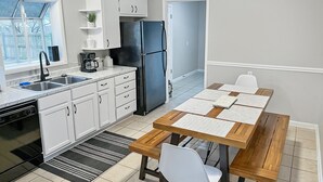 A modern kitchen with white cabinets, a black refrigerator, a coffee maker, and a dining area with a wooden table and white chairs. - Red Tail Cottage: Full Fence, Hammock Chair, Walk to Bass Pro, Movie Night (Springfield)