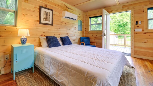 A wooden cabin bedroom with a bed, a nightstand, an air conditioning unit, and a window. - Cozy Hiker's Hideaway: Steps From Creeper Trail! (Damascus)
