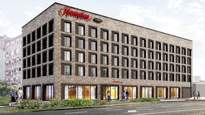 Un bâtiment moderne à la façade en briques, doté de nombreuses fenêtres et d’une enseigne « Hampton by Hilton » bien visible.