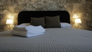 A neatly made bed with folded white towels, a textured white bedspread, and a dark headboard. There are two bedside lamps with warm light, and the wall features a leaf-patterned wallpaper. - Sikelìa Bed & Breakfast (Carini)