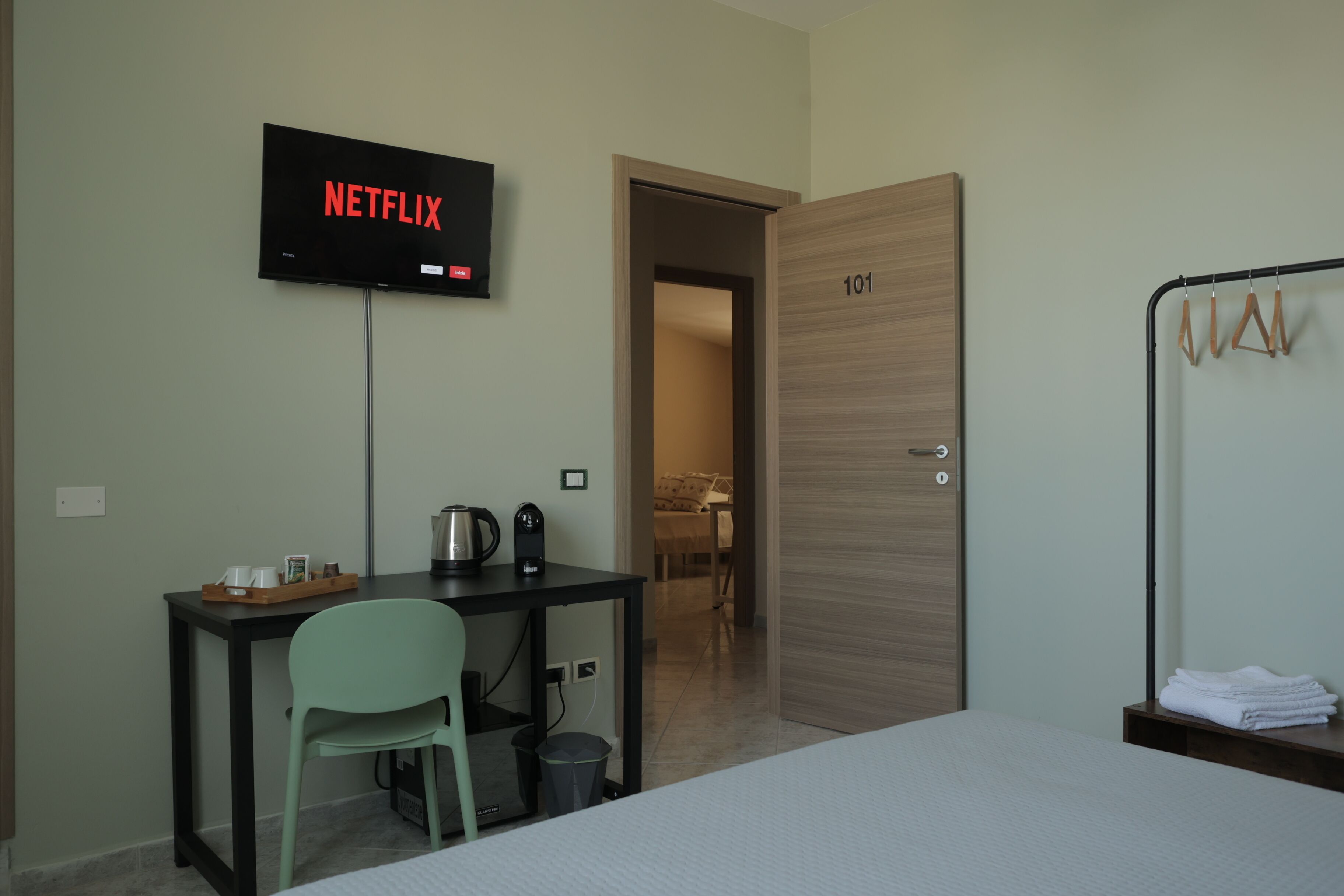 A hotel room with a bed, a desk with a chair, a television displaying 'NETFLIX', a door labeled '101', and a coat rack with hangers.