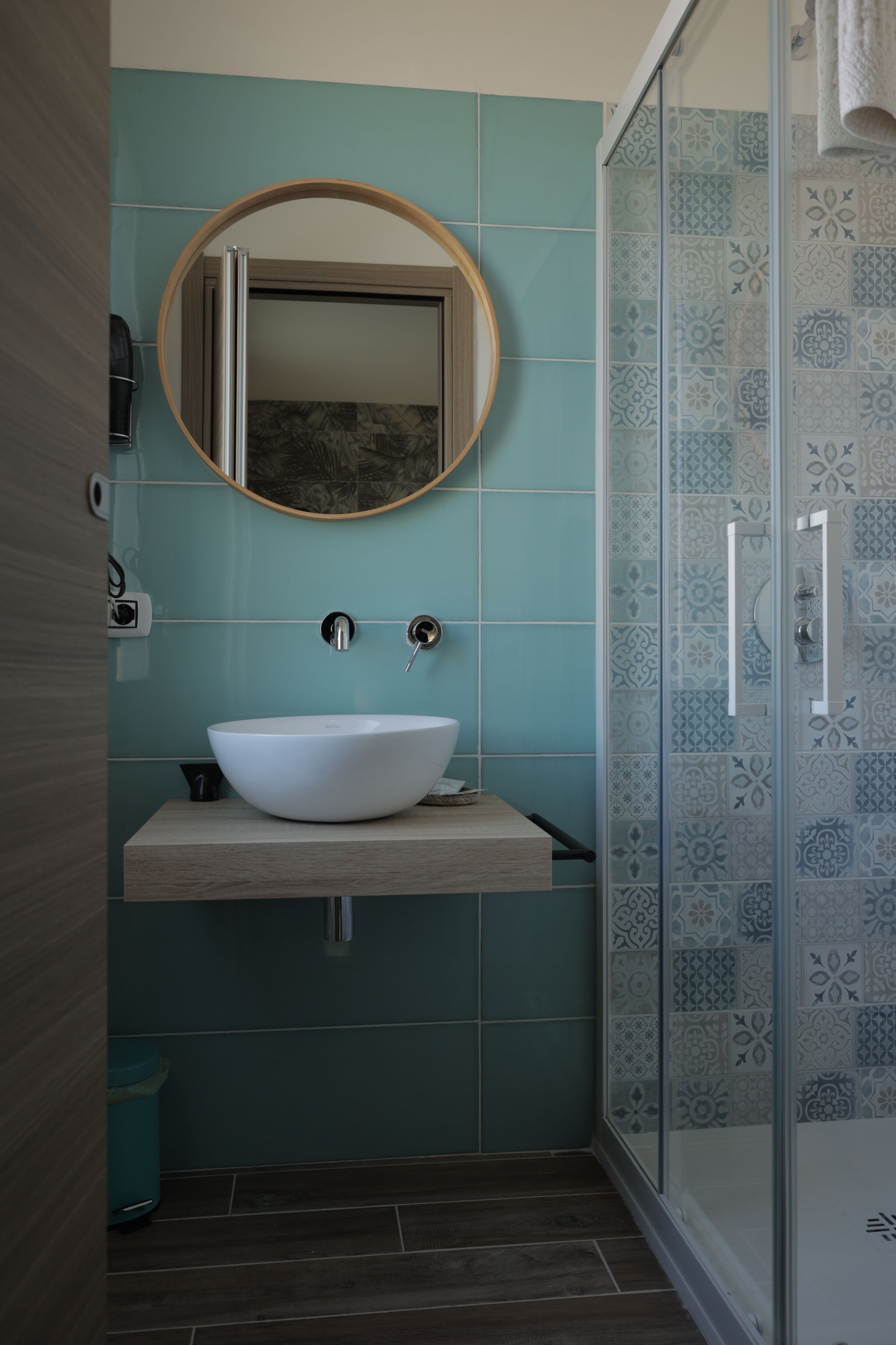 Un baño con lavamanos blanco, un espejo redondo y una ducha con azulejos estampados.