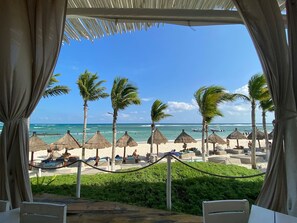 A beach view from a covered area with palm trees and thatched umbrellas.