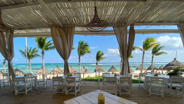 Zona salotto esterna coperta con mobili bianchi e un tavolo, affacciata su una spiaggia con palme e l'oceano.