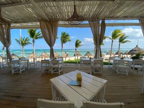 A covered outdoor seating area with white furniture and a table, overlooking a beach with palm trees and the ocean.