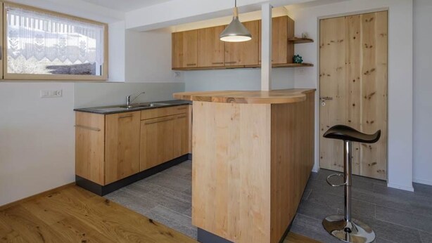 A modern kitchen with wooden cabinets, a central island, a bar stool, and a window with sheer curtains. - Guarda Val Müller Erlenstube by Interhome (Lantsch)