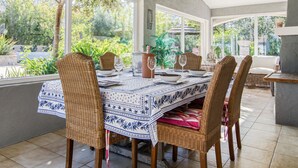 Een eetruimte met rieten stoelen en een tafel voor vier personen, grote ramen met uitzicht op groen buiten.
