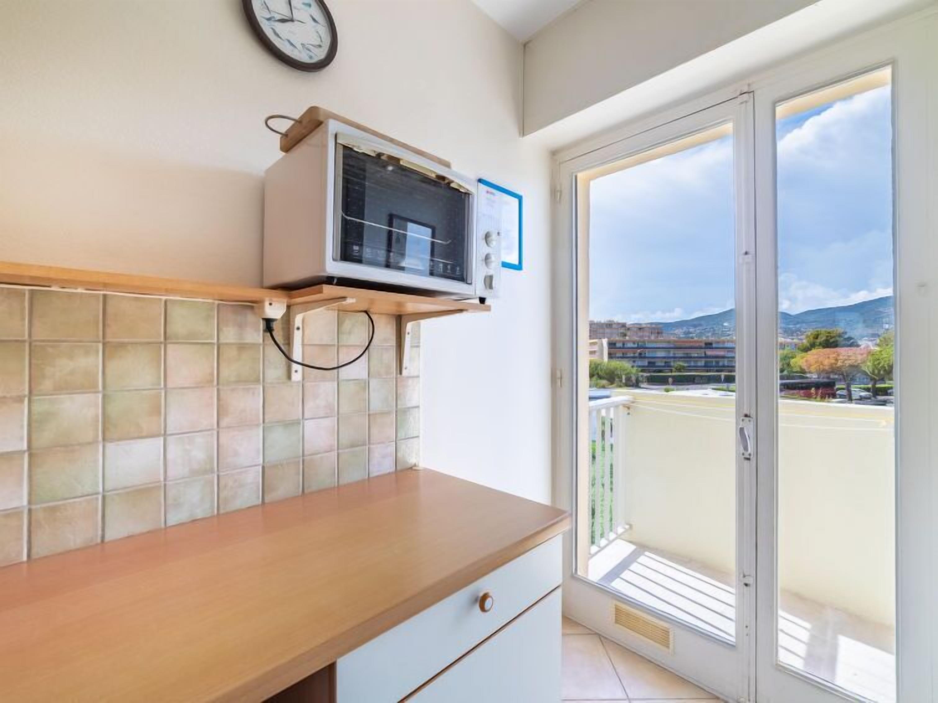 A kitchen with a microwave, a clock, and a view of the outside.