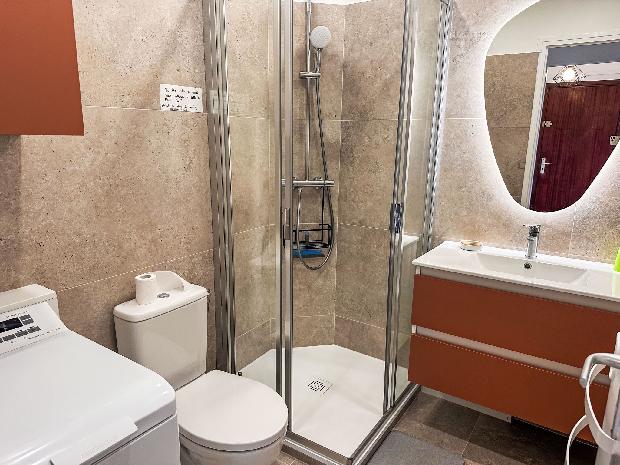 A modern bathroom with a glass-enclosed shower, a toilet, a sink with a mirror, and a washing machine.