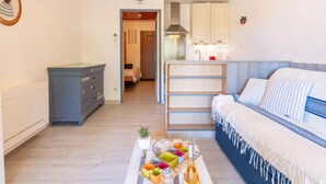 Un soggiorno moderno con un divano, un tavolino da caffè con oggetti colorati, una ventola a soffitto e una cucina visibile attraverso una porta aperta.