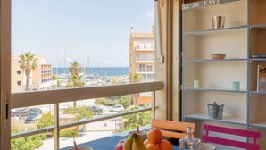 Un balcone con vista su un porto turistico e degli edifici, un tavolo con frutta e una ciotola, e uno scaffale con oggetti decorativi.