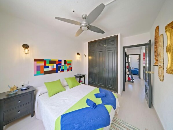Une chambre à coucher comprenant un lit, un ventilateur de plafond, un tableau décoratif mural, une armoire et une porte donnant sur une autre pièce.
