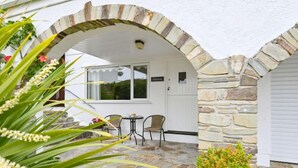 Un arco in pietra conduce a un portico coperto con tavolo e sedie, una porta con l'indicazione 'Edyreen' e un piccolo arbusto sullo sfondo.