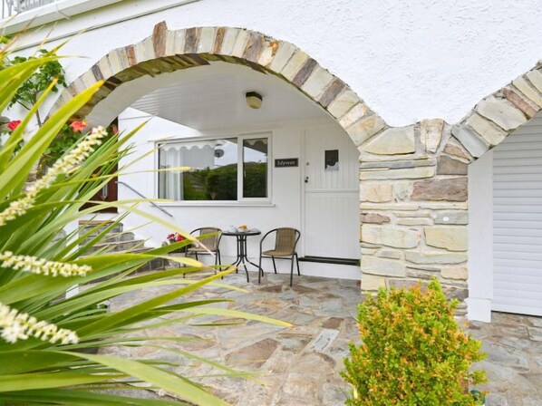 A stone archway leading to a covered patio area with a table and chairs, a door labeled 'Edyreen', and a small bush in the foreground.