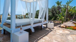 A white canopy bed with a wooden deck, surrounded by tropical plants and a stone pathway. - Sa Caseta by Interhome (Ses Salines)