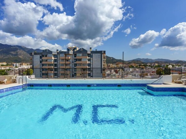 Ein Schwimmbecken mit dem Schriftzug 'MC' im Wasser, umgeben von einem modernen Gebäude mit Balkonen, unter blauem Himmel.