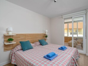 Ein Schlafzimmer mit einem Holzkopfteil, zwei Nachttischen mit Lampen, einem Bett mit gemusterten Bettbezügen und Blick durch eine Schiebetür auf einen Außenbereich.