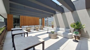 An outdoor seating area with tables, chairs, and a sofa, surrounded by a modern building with large windows and a striped awning. - Zahle Jardins (São Paulo)