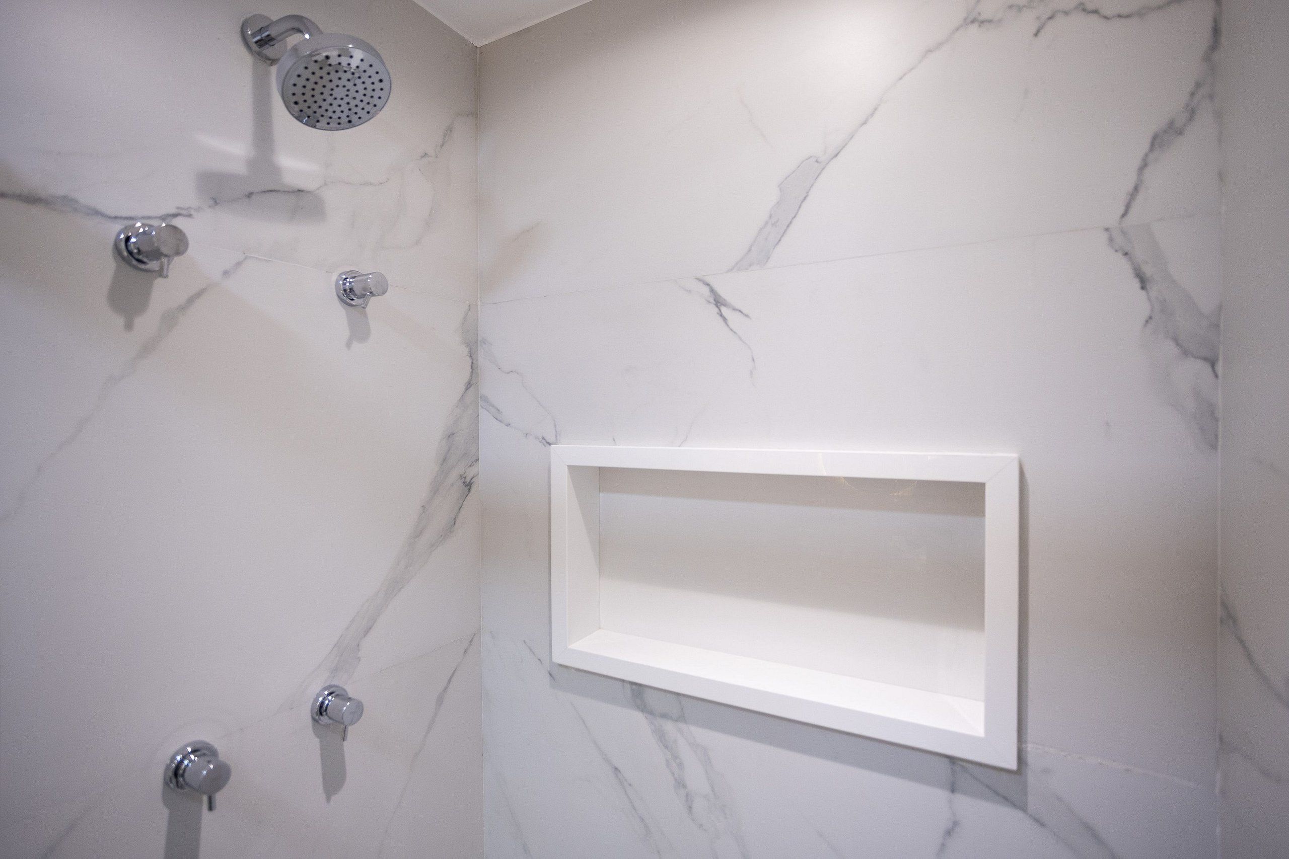 Un baño con pared de mármol, un cabezal de ducha y una repisa.