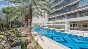 A swimming pool with geometric tile patterns, surrounded by lounge chairs and palm trees, adjacent to a modern building with balconies.