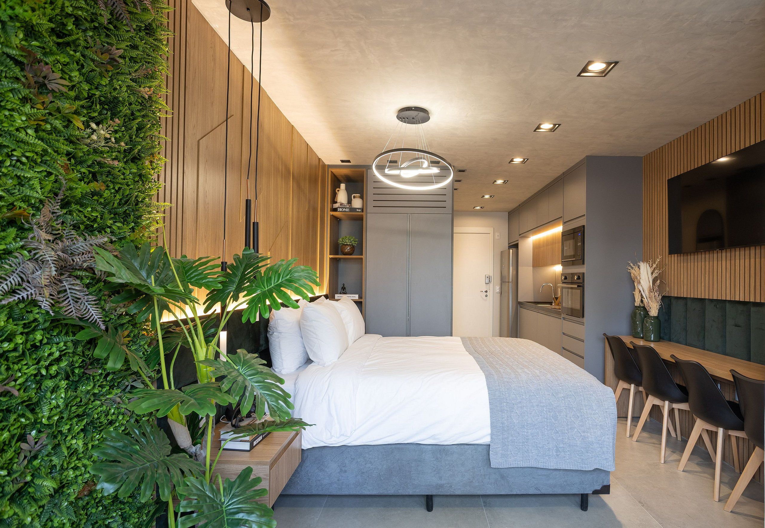 Ein modernes Schlafzimmer mit einem Bett, Pflanzen auf dem Nachttisch, einer Holzwand und einem Essbereich mit Stühlen.
