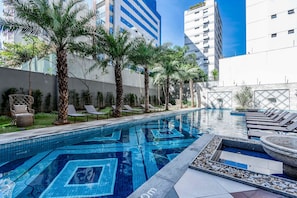 Una piscina con diseños de mosaicos, sillas de descanso, palmeras y edificios modernos al fondo.