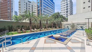 A swimming pool with multiple lanes, surrounded by lounge chairs and palm trees, with tall buildings in the background. - Zahle Jardins (São Paulo)