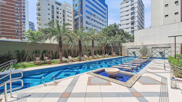 A swimming pool with multiple lanes, surrounded by lounge chairs and palm trees, with tall buildings in the background.