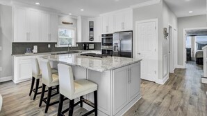 A modern kitchen with white cabinetry, a large island with bar stools, stainless steel appliances, and a grey backsplash. - Grand Pavilion 34 by Wild Dunes, Luxury Home (Isle of Palms)