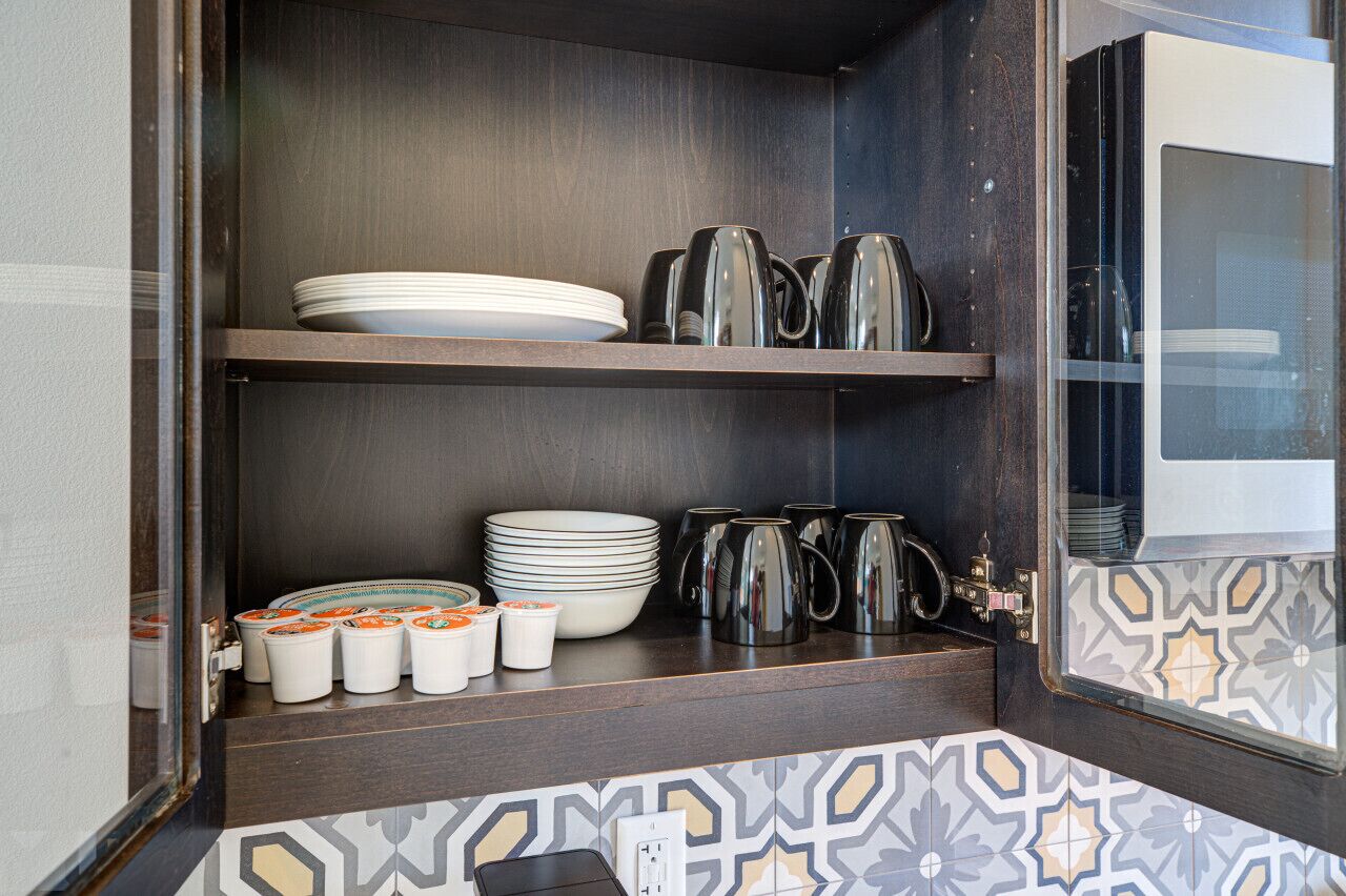 A compact kitchenette with dark wood shelves, stacked plates, mugs, and a coffee maker.