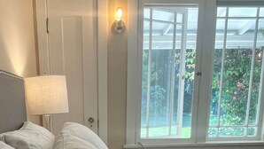 Een slaapkamer met een bed, kussens, een nachtkastje, een lamp en een raam met uitzicht op groen.