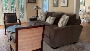 Een woonkamer met een bruine bank, een rieten stoel, een kleine tafel en een kamerkleed met een patroon. Er zijn twee ramen met gebogen bovenkanten en een deur die naar buiten leidt.