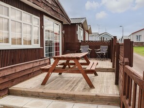 A wooden deck with a picnic table and two chairs, adjacent to a house with large windows and a wooden fence.