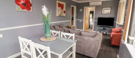Ein modernes Wohnzimmer mit Esstisch, weißen Stühlen, einem grauen Sofa und einem Fernseher.