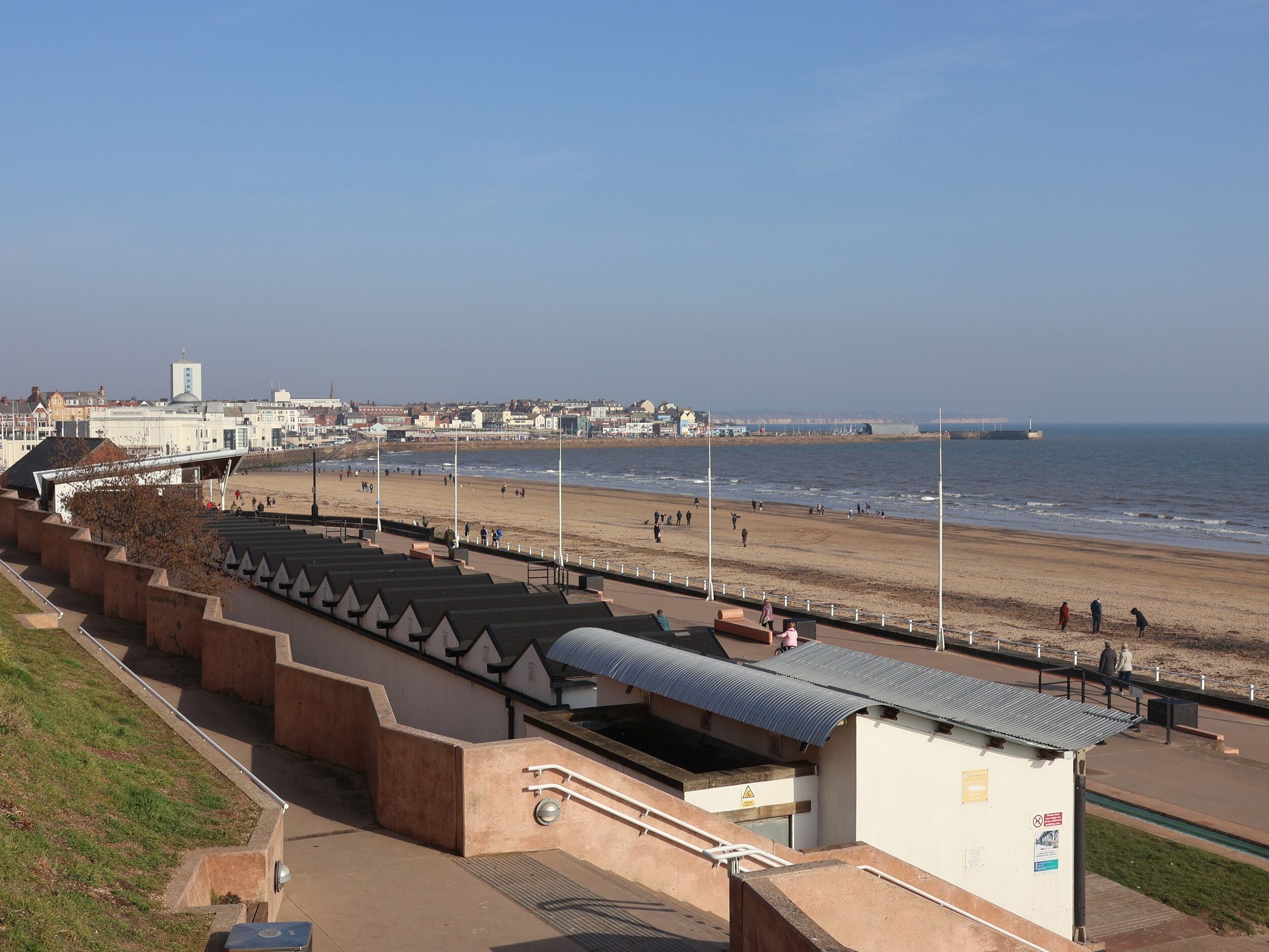 A coastal promenade with a series of beach huts, a promenade with a metal roof, and a view of the sea and buildings in the distance.