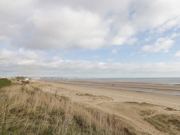 A wide sandy beach with gentle waves, grassy dunes, and a distant shoreline with buildings.