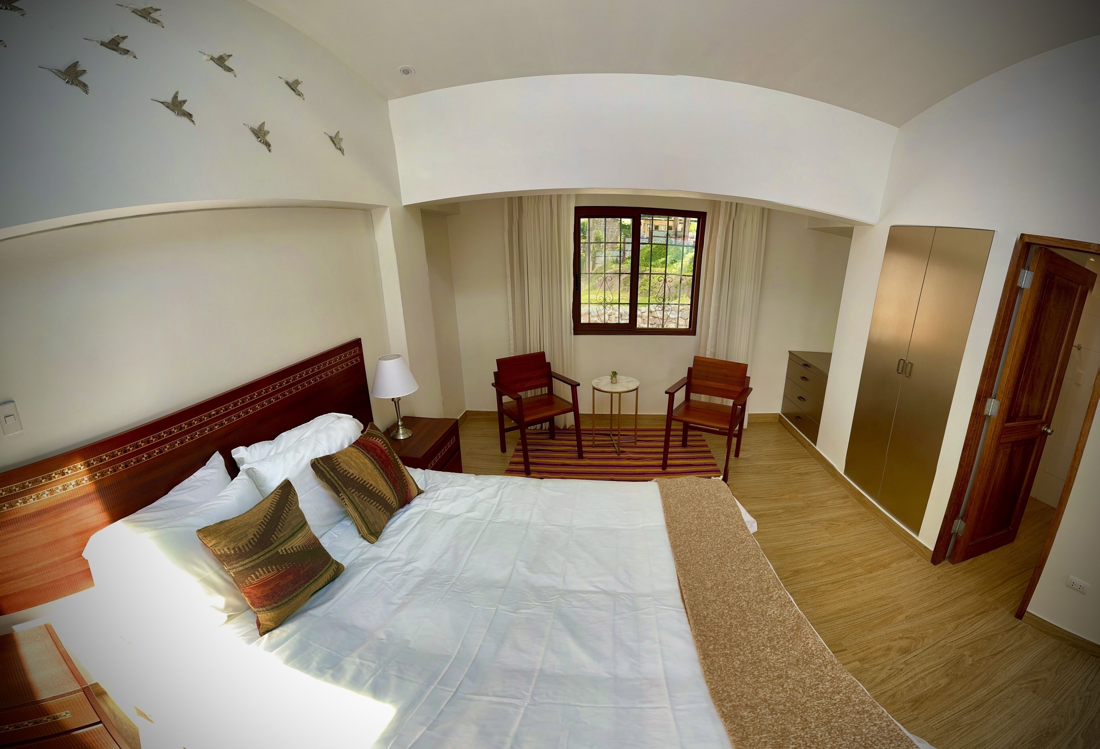 A hotel room with a large bed, a desk, two chairs, a window with curtains, and a wardrobe.