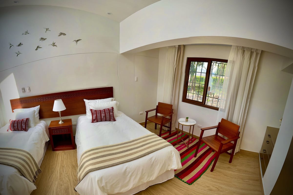 Habitación de hotel con dos camas, cabecero de madera, una mesita con sillas, una ventana con cortinas y un adorno de pájaro en la pared.