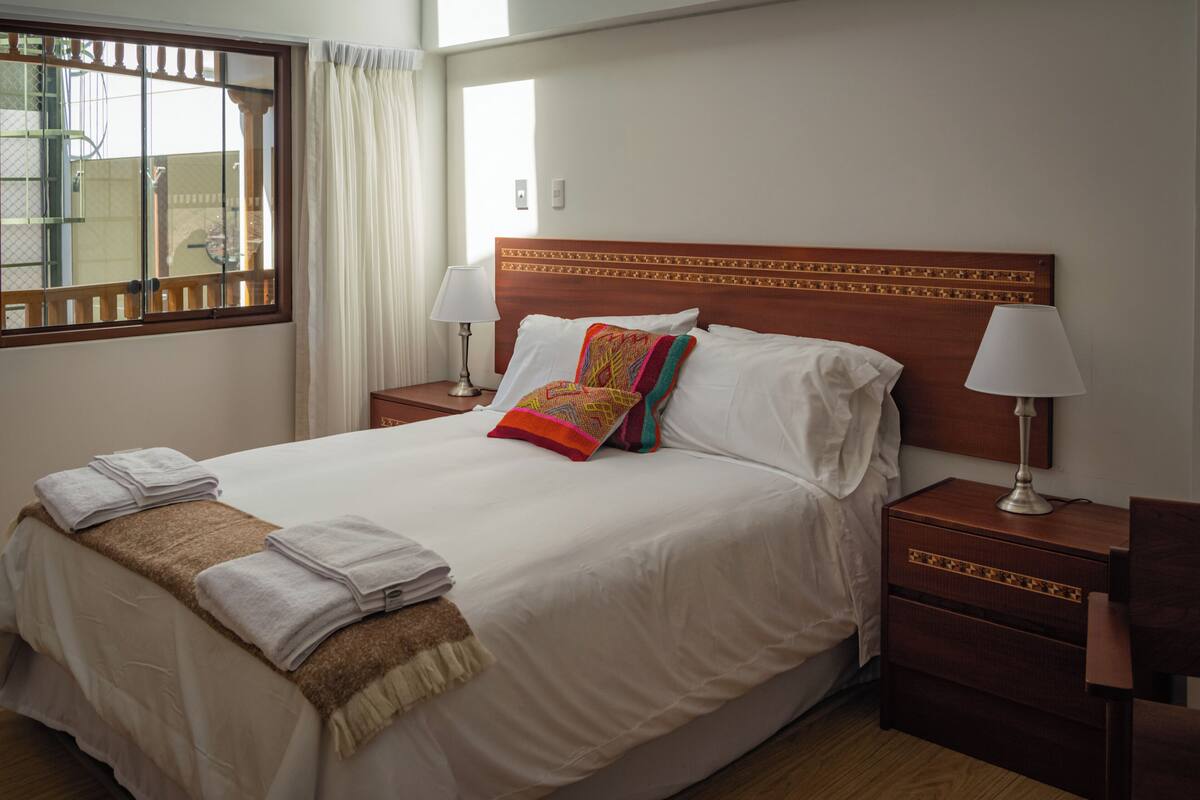 Una cama bien hecha con sábanas blancas, un cojín decorativo de colores y una toalla doblada a un lado. Hay dos mesitas de noche con lámparas y una ventana con cortinas.